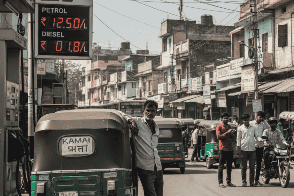 A realistic street scene in a small city showing an auto-rickshaw driver leaning pensively against his vehicle at a petrol station. In the background, a digital sign displays rising fuel prices, while other commuters and vehicles navigate a busy, dusty road, illustrating the economic pressure on small transport operators.