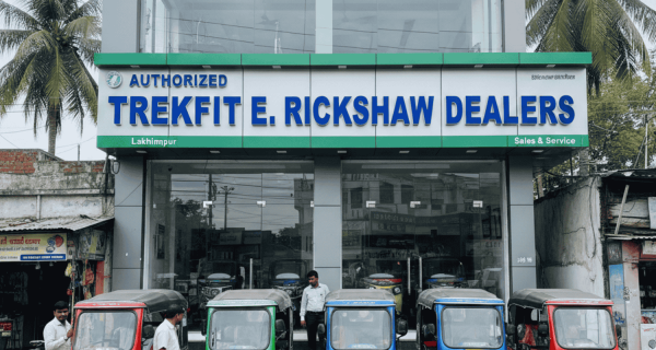 A modern two-story Trekfit E-Rickshaw showroom in Lakhimpur featuring a large glass storefront. Several colorful e-rickshaws in red, green, and blue are parked in front, with a prominent sign reading