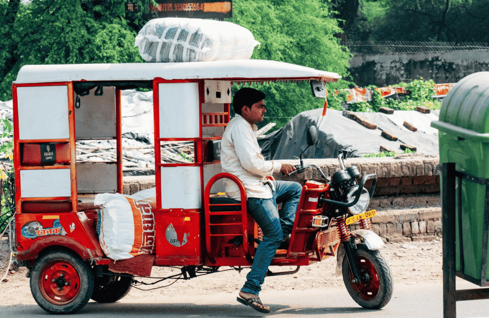 A side-view showing a red and white electric rickshaw (e-rickshaw) driving along a street. The driver, a man wearing a white shirt and blue jeans, is seated at the handlebars. A large, white woven sack is strapped securely to the roof of the vehicle, and another large sack is visible inside the passenger compartment. A green waste bin is partially visible in the right foreground, with trees and a brick wall in the background.